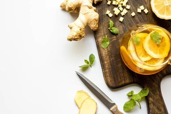 Vue du dessus du thé chaud près de la racine de gingembre, citron et menthe sur planche à découper en bois avec couteau sur fond blanc — Photo de stock