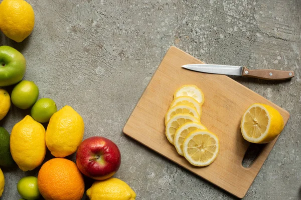 Top view of tasty colorful fruits and wooden cutting board with lemon slices and knife on concrete grey surface — Stock Photo