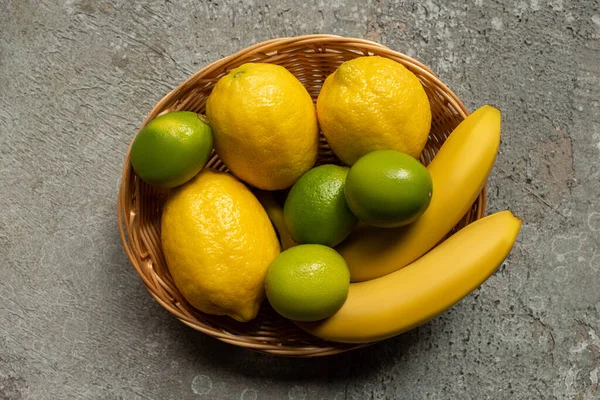 Top view of colorful bananas, limes and lemons in wicker basket on grey concrete surface — Stock Photo