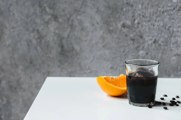 Холодный кофе со льдом в стакане возле апельсина и кофейных зерен на белом столе — стоковое фото