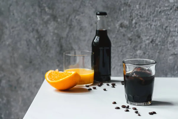 Foco seletivo de café frio brew com gelo em vidro e garrafa perto de suco de laranja e grãos de café na mesa branca — Fotografia de Stock