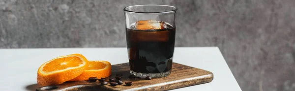 Холодный кофе со льдом в стакане возле оранжевых ломтиков на доске и кофейных зерен на белом столе, панорамный снимок — стоковое фото