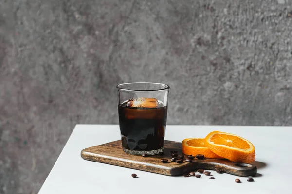 Café de cerveza fría con hielo en vidrio cerca de rebanadas de naranja en la tabla de cortar y granos de café en la mesa blanca - foto de stock