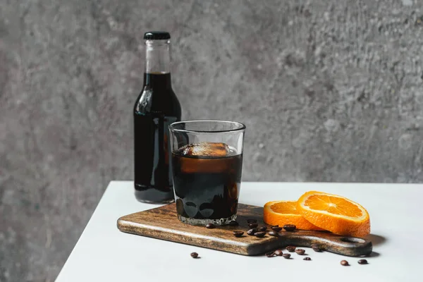 Café de cerveza fría con hielo en vidrio y botella cerca de rebanadas de naranja en la tabla de cortar y granos de café en la mesa blanca - foto de stock
