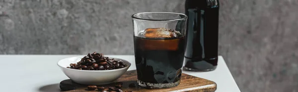 Cold brew coffee with ice in glass and bottle near coffee beans on chopping board on white table, panoramic shot — Stock Photo