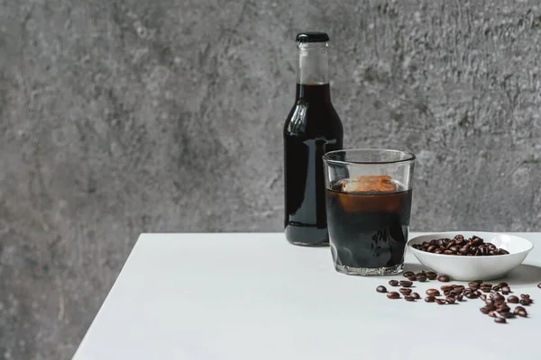 Café de cerveza fría con hielo en vidrio y botella cerca de granos de café en la mesa blanca - foto de stock