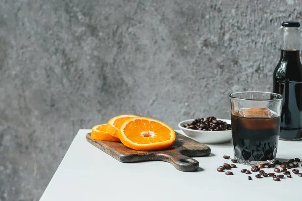 Café froid infusé avec glace en verre et bouteille près de tranches d'orange sur planche à découper et grains de café sur table blanche — Photo de stock