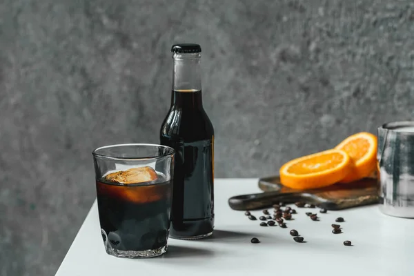 Selective focus of cold brew coffee with ice in glass and bottle near orange slices on chopping board and coffee beans on white table — Stock Photo