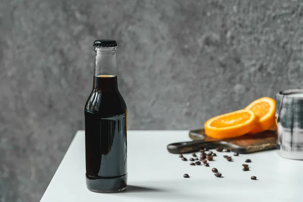 Messa a fuoco selettiva di caffè freddo birra con ghiaccio in bottiglia vicino fette di arancia sul tagliere e chicchi di caffè sul tavolo bianco — Foto stock