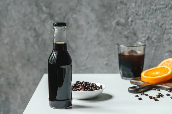 Foco seletivo de café frio brew com gelo em garrafa e vidro perto de fatias de laranja na tábua de corte e grãos de café na mesa branca — Stock Photo