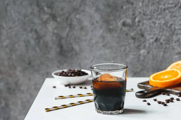 Selektiver Fokus von kaltem Brühkaffee mit Eis im Glas in der Nähe von Orangenscheiben auf Schneidebrett, Trinkhalmen, Kaffeebohnen auf weißem Tisch — Stockfoto