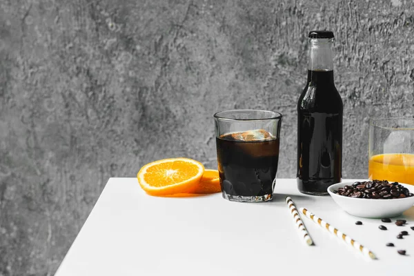 Messa a fuoco selettiva del caffè freddo con ghiaccio in vetro e bottiglia vicino al succo d'arancia, cannucce e chicchi di caffè sul tavolo bianco — Foto stock