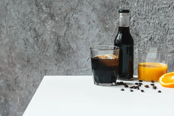 Холодный кофе со льдом в стакане и бутылка возле апельсинового сока и кофейных зерен на белом столе — стоковое фото
