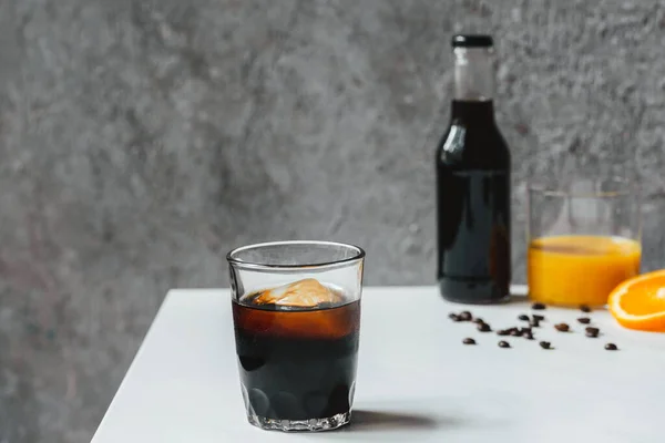 Selektiver Fokus von kaltem Brühkaffee mit Eis im Glas und Flasche in der Nähe von Orangensaft und Kaffeebohnen auf weißem Tisch — Stockfoto