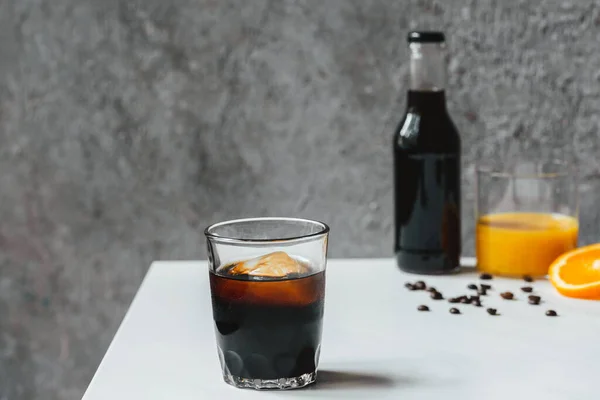 Selective focus of cold brew coffee with ice in glass and bottle near orange juice and coffee beans on white table — Stock Photo