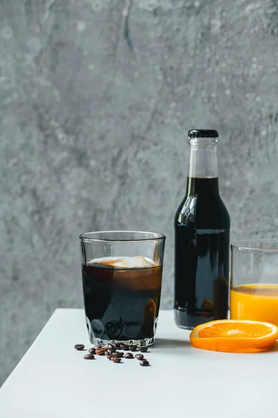 Café de cerveza fría con hielo en vidrio y botella cerca del jugo de naranja y granos de café en la mesa blanca - foto de stock