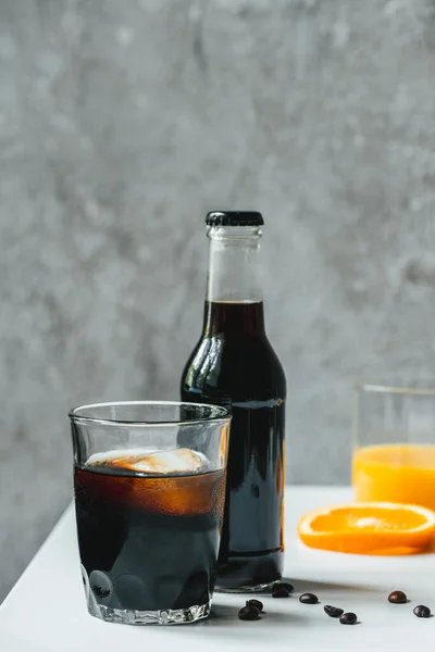 Selective focus of cold brew coffee with ice in glass and bottle near orange juice and coffee beans on white table — Stock Photo