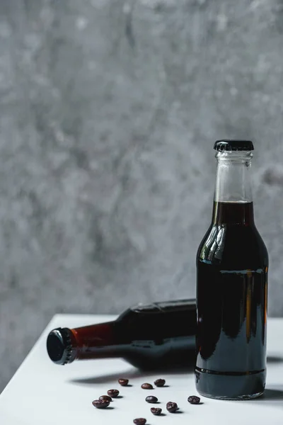 Холодный кофе в бутылках возле кофейных зерен на белом столе — стоковое фото