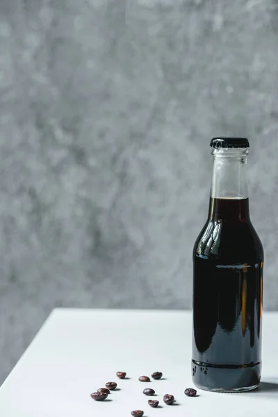 Холодный кофе в бутылке возле кофейных зерен на белом столе — стоковое фото