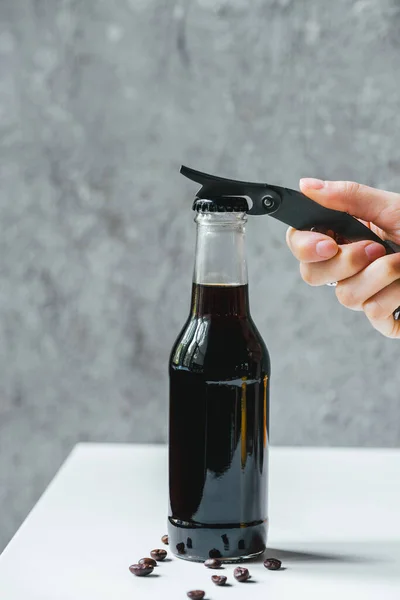 Vista recortada de la mujer abriendo café de cerveza fría en botella con abridor en mesa blanca con granos de café - foto de stock