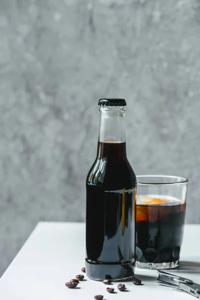 Café infusé froid avec glace en verre et bouteille près des grains de café et ouvre-table sur table blanche — Photo de stock