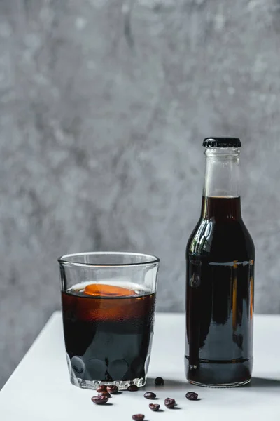 Холодный кофе со льдом в стекле и бутылка возле кофейных зерен на белом столе — стоковое фото