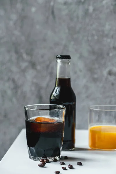 Messa a fuoco selettiva di caffè freddo birra con ghiaccio in vetro e bottiglia vicino succo d'arancia e chicchi di caffè sul tavolo bianco — Foto stock