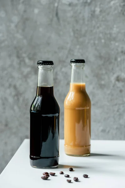 Foco seletivo de café cerveja fria em garrafas perto de grãos de café na mesa branca — Fotografia de Stock