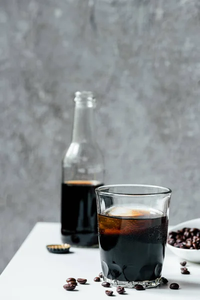 Enfoque selectivo de café de cerveza fría con hielo en vidrio cerca de la botella y granos de café en la mesa blanca - foto de stock
