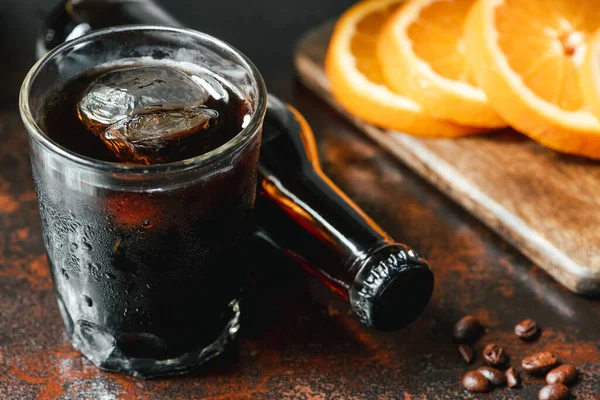 Selective focus of cold brew coffee with ice in glass and bottle near orange slices and coffee beans on rusty surface — Stock Photo