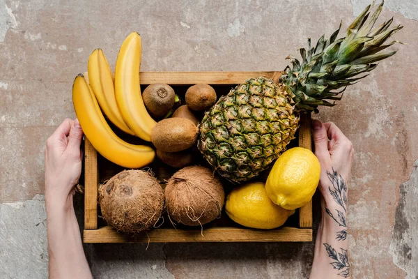 Vista ritagliata della donna che tiene i frutti tropicali maturi in scatola di legno sulla superficie alterata — Foto stock