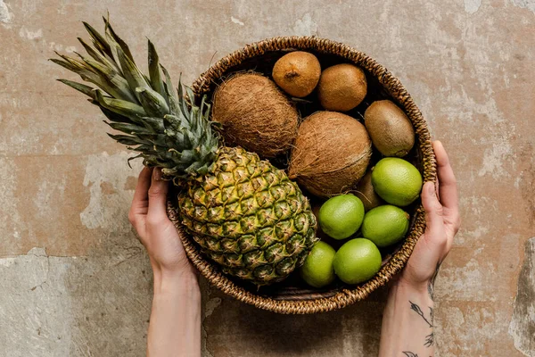Cropped view of woman holding ripe exotic fruits in wicker basket on weathered surface — Stock Photo