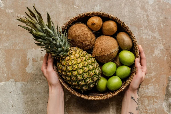 Vista recortada de la mujer sosteniendo frutas exóticas maduras en canasta de mimbre en la superficie erosionada - foto de stock