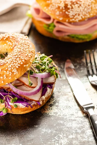 Selective focus of fresh delicious bagel with meat, red onion, cream cheese and sprouts near cutlery on textured surface — Stock Photo
