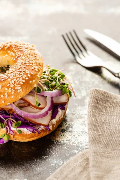 Foco seletivo de bagel delicioso fresco com carne, cebola vermelha, queijo creme e brotos perto de guardanapo com talheres na superfície texturizada — Fotografia de Stock