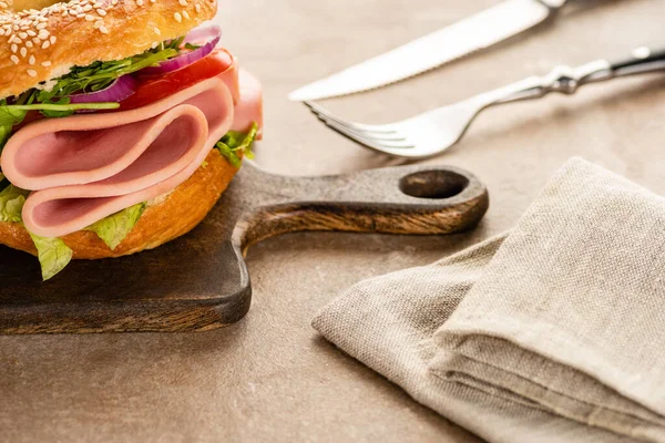 Selective focus of fresh delicious bagel with sausage on wooden cutting board near napkin and cutlery on textured surface — Stock Photo