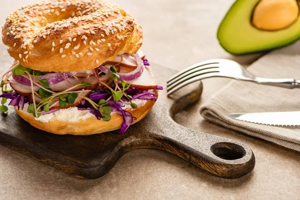 Fresh delicious bagel with meat, red onion, cream cheese and sprouts on wooden cutting board near napkin and cutlery — Stock Photo