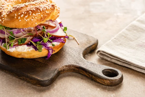 Delizioso bagel fresco con carne, cipolla rossa, crema di formaggio e germogli sul tagliere di legno vicino al tovagliolo — Foto stock