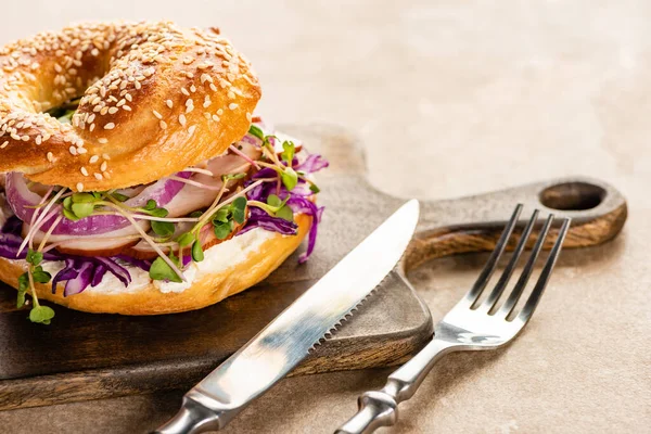 Bagel delicioso fresco com carne, cebola vermelha, queijo creme e brotos em tábua de corte de madeira com talheres — Fotografia de Stock