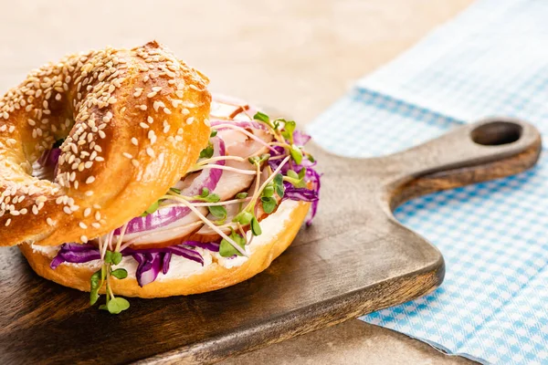 Frischer leckerer Bagel mit Fleisch, roten Zwiebeln, Frischkäse und Rosenkohl auf Holzschneidebrett in der Nähe karierter Serviette — Stockfoto