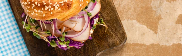 Top view of fresh delicious bagel with meat, red onion, cream cheese and sprouts on wooden cutting board and plaid blue napkin, panoramic shot — Stock Photo