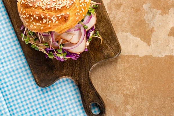 Top view of fresh delicious bagel with meat, red onion, cream cheese and sprouts on wooden cutting board and plaid blue napkin — Stock Photo