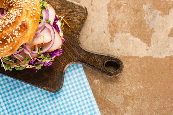 Top view of fresh delicious bagel with meat, red onion, cream cheese and sprouts on wooden cutting board and plaid blue napkin — Stock Photo