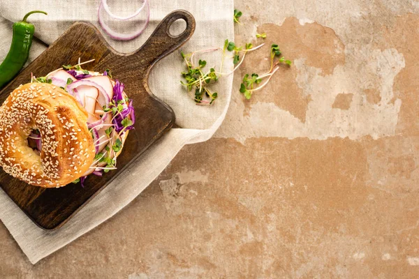 Top view of fresh delicious bagel with meat, red onion and sprouts on wooden cutting board on napkin with jalapenos on aged beige surface — Stock Photo