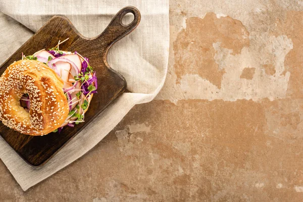 Top view of fresh delicious bagel with meat, red onion and sprouts on wooden cutting board on napkin on aged beige surface — Stock Photo