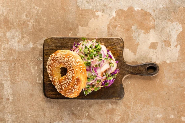 Top view of fresh delicious bagel with meat, red onion and sprouts on wooden cutting board on aged beige surface — Stock Photo