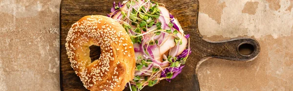 Top view of fresh delicious bagel with meat, red onion and sprouts on wooden cutting board on aged beige surface, panoramic shot — Stock Photo