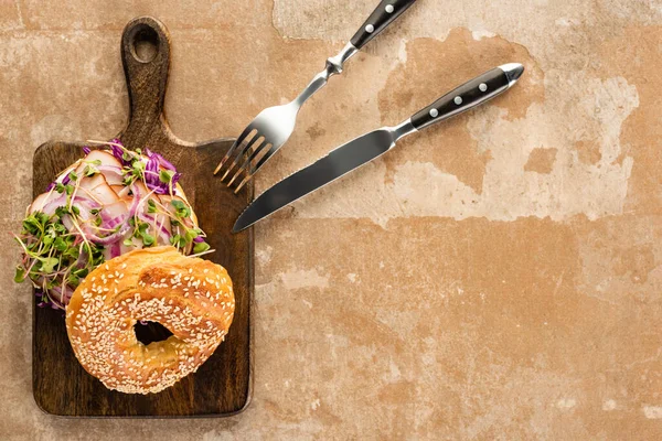 Top view of fresh delicious bagel with meat, red onion and sprouts on wooden cutting board with cutlery on aged beige surface — Stock Photo