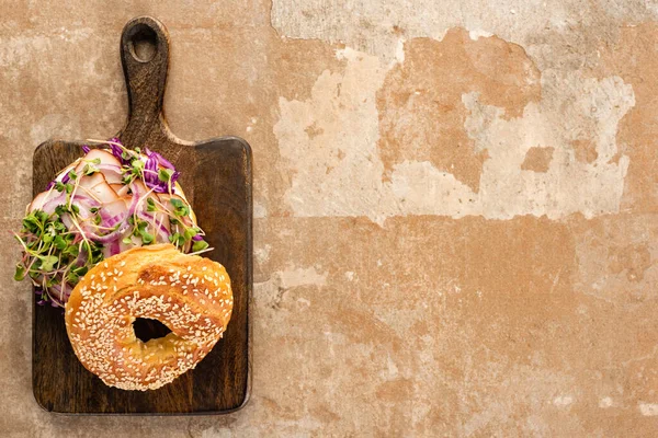 Vista superior de bagel delicioso fresco com carne, cebola vermelha e brotos na placa de corte de madeira na superfície bege envelhecida — Fotografia de Stock