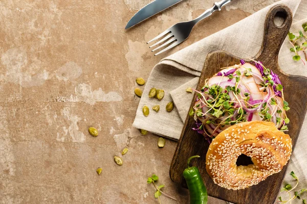 Vista superior de bagel delicioso fresco com carne, cebola vermelha e brotos em tábua de corte de madeira perto de guardanapo e talheres na superfície bege envelhecida — Fotografia de Stock